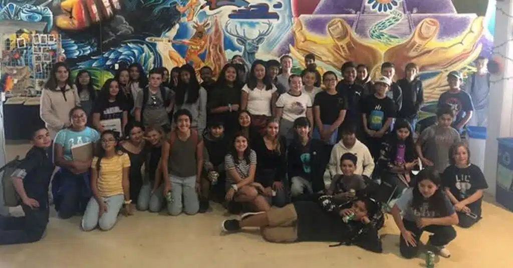 A group of people with two-toned hair posing indoors against a vibrant, colorful mural featuring abstract designs and hands. Most are smiling, exuding a friendly and lively atmosphere.