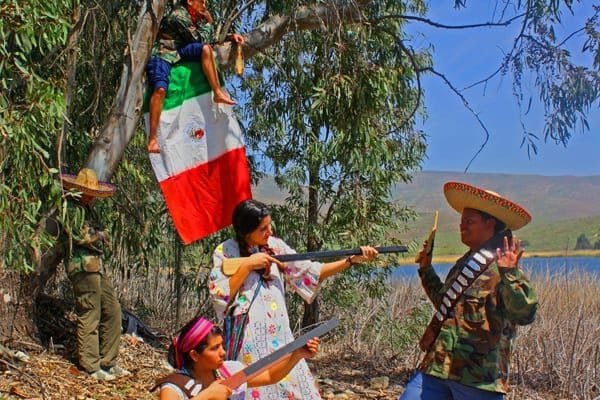 A group of people in colorful attire and camouflage, posing outdoors with toy guns and a large flag, next to a tree. The setting is a grassy area near water with hills in the background, under a clear blue sky.