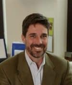 A person with short dark hair and a beard is smiling while wearing a brown blazer and a white shirt. Seated indoors, surrounded by books and a framed picture of a plant illustration in the background, they exude an air of warmth and wisdom.