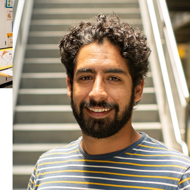 A smiling man with curly hair and a beard is wearing a striped shirt. He is standing in front of a blurred staircase.