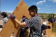 People working outdoors, handling large wooden panels under a clear sky. A man in the foreground wears a striped shirt and gloves, assisting with the construction work. Others in the background are also engaged in similar tasks.