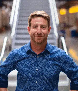 Randy Scherer stands confidently in a blue patterned shirt, arms crossed and smiling warmly. He is positioned before a blurred background that features an illuminated staircase, creating a scene with a casual and approachable atmosphere.