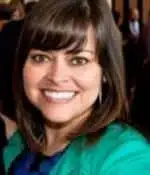 A woman with shoulder-length brown hair and bangs smiles at the camera, her green jacket draped over a dark top. As if narrating her own life story, the blurred background features people in formal attire, adding an intriguing contrast to the scene.