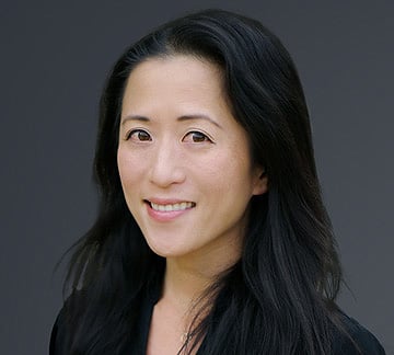 A person with long, dark hair smiles softly against a dark background, wearing a black top.