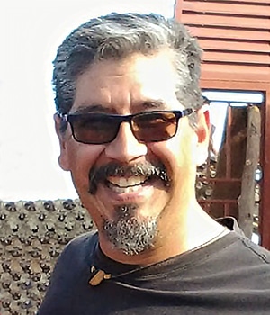 A man with short gray hair, a mustache, and a goatee is smiling like a guest on his favorite episode, wearing sunglasses and a black shirt. He stands outdoors against a backdrop featuring a wooden structure and textured wall.