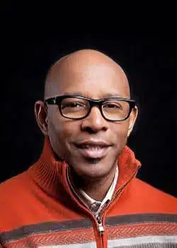 A person wearing black glasses and an orange sweater with a zip-up collar, smiling as if ready to start a podcast, stands against a black background.