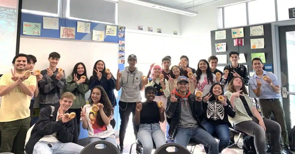 A group of students in a classroom holding donuts and smiling. They are standing and sitting, with colorful artwork on the walls and natural light from windows. Some students are making a W with their hands.