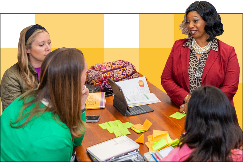 A group of four people sits around a table covered with a notebook, colorful sticky notes, and a tablet. One person in a red jacket stands speaking about deeper learning, while the others listen attentively. In the background, a yellow checkerboard pattern adds vibrancy to their discussion.