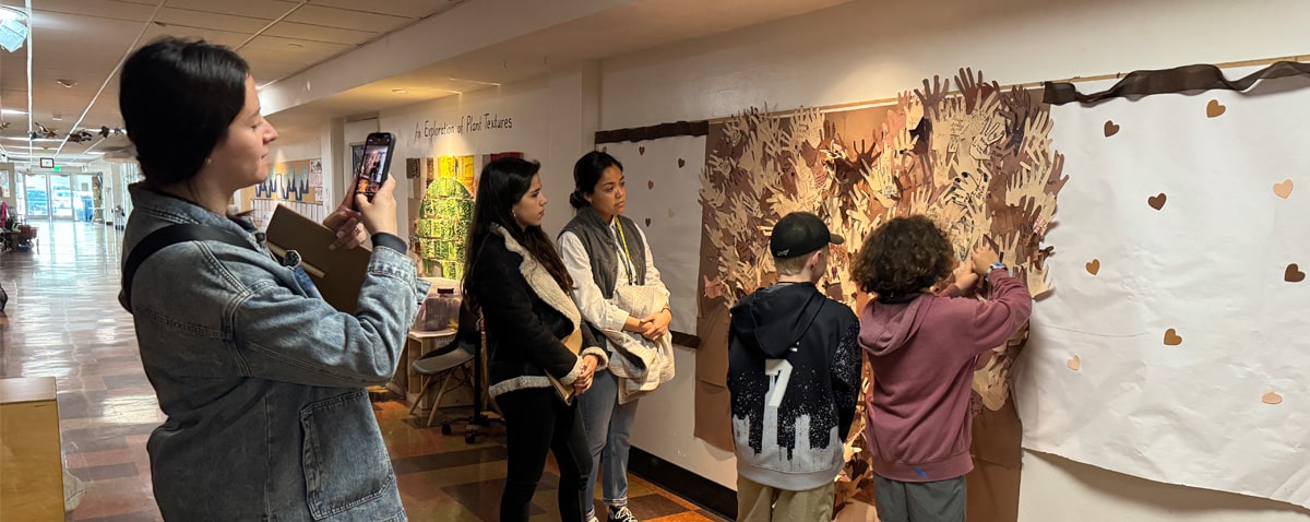 During an HTH School Visits event on Jan 16, 2025, people admire a wall art display with paper hand cutouts in a hallway. One person captures the moment on their smartphone while two children engage with the artwork. Informational posters and a table with activities complete the scene.