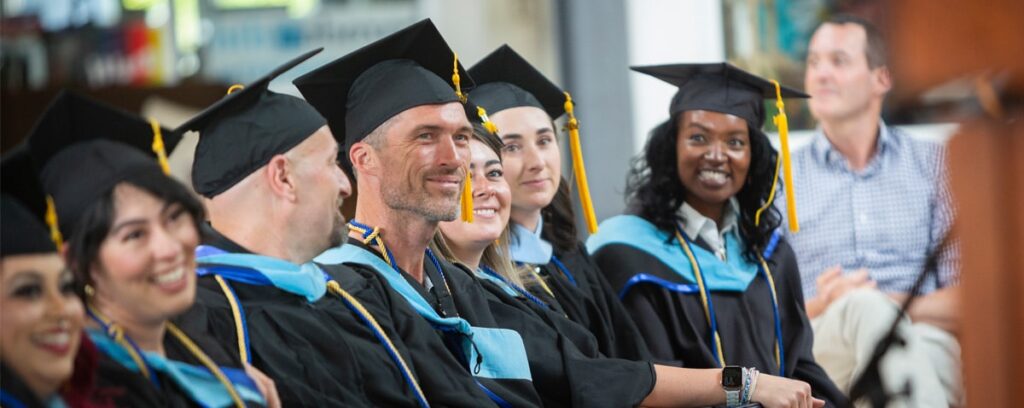 A group of smiling graduates in caps and gowns sit together, facing forward at their graduation ceremony. Members of this Masters Program showcase tassels on their caps. A person in casual clothes is seated behind them, perhaps contemplating an educational leadership journey.