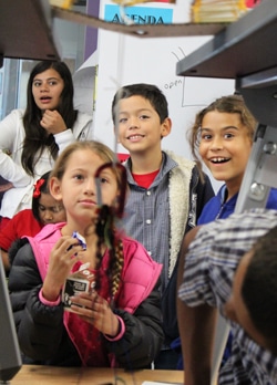 A group of children, including a girl in a pink jacket and a boy in a red shirt, are gathered around a science experiment in a classroom. They are looking intently at the demonstration, showing curiosity and excitement.