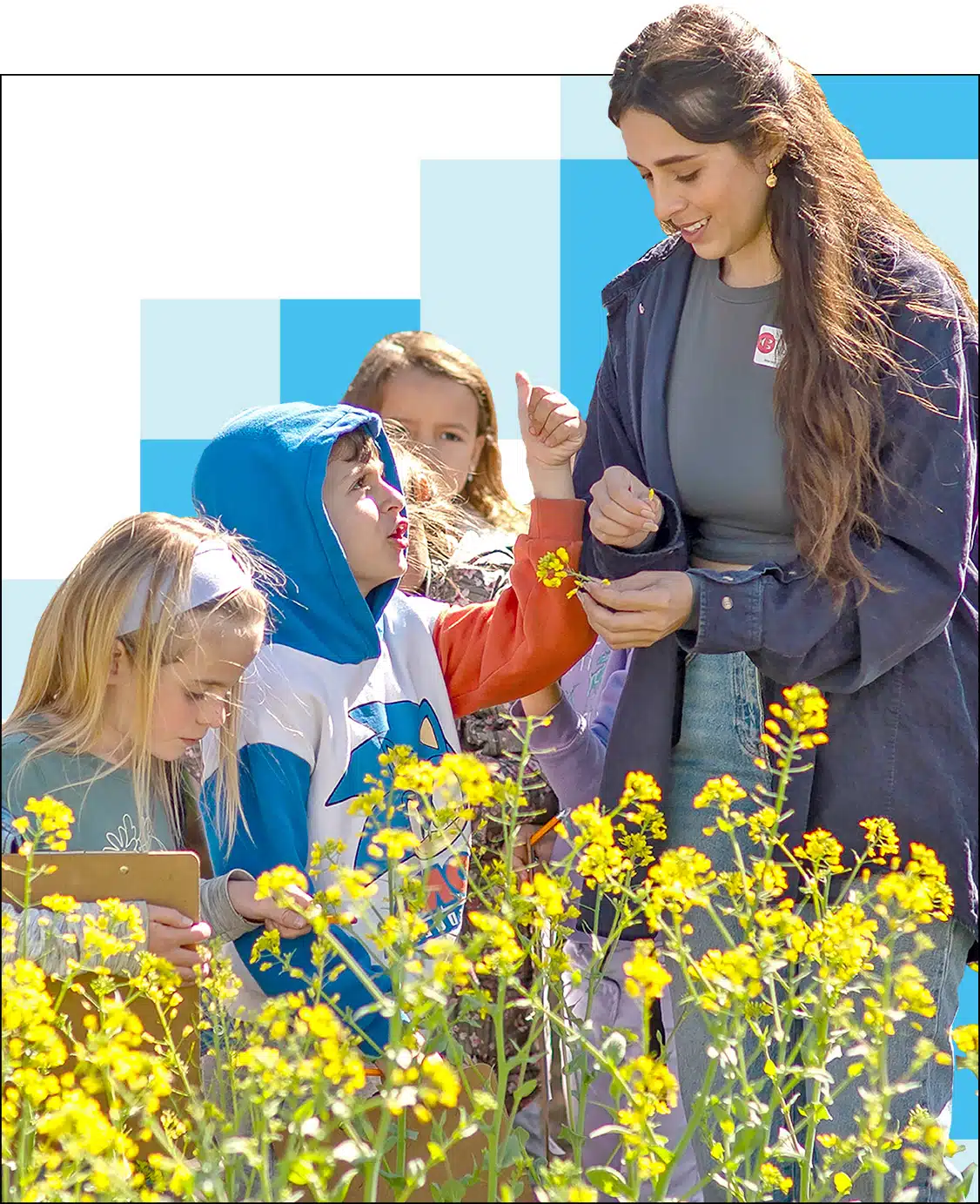 A woman from the Hi Tech High Graduate School of Education is smiling while showing yellow flowers to a group of three children outdoors. The children are observing and appear engaged, with one child pointing at the flowers. The scene is bright and cheerful.
