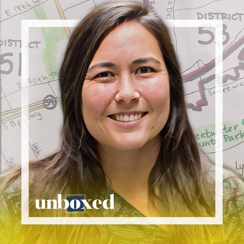 A person with long hair smiles at the camera, embodying the spirit of electioneering. The background showcases a map of district numbers and street names, hinting at recent redistricting changes. The word unboxed is visible at the bottom.