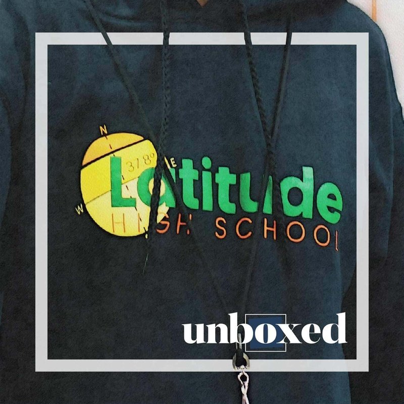 A person wearing a dark hoodie with Latitude High School in green and orange text, featuring a yellow globe design framed by a white border. The word unboxed, emblematic of what matters most during lockdown, is printed at the bottom.