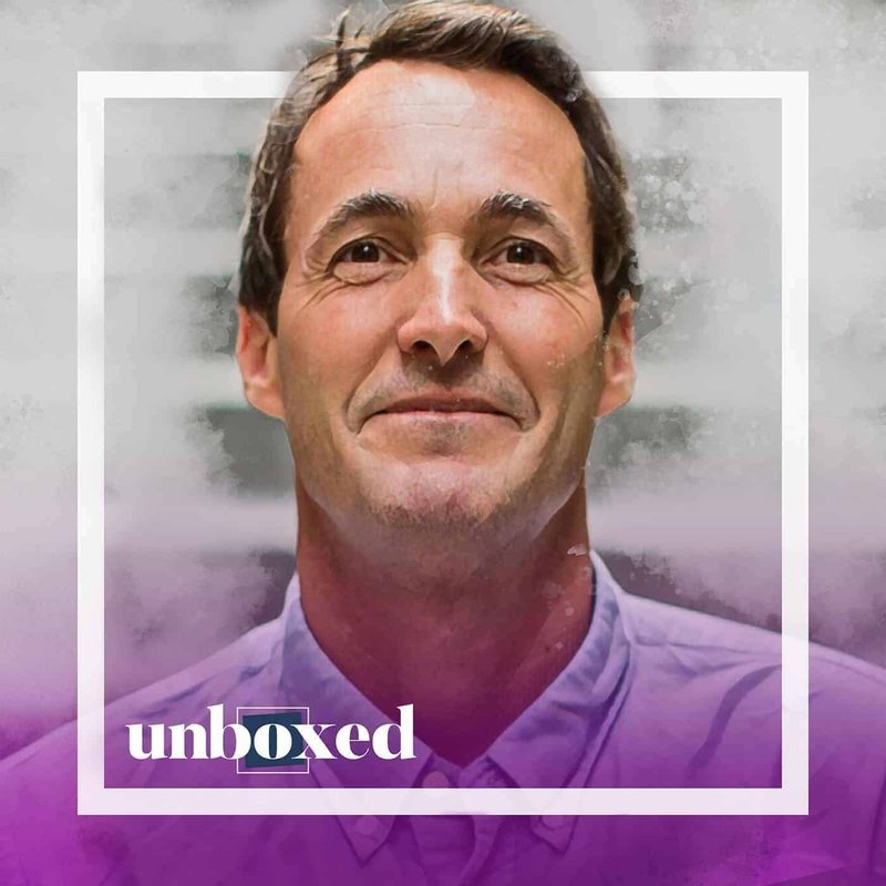 John Santos, a man with short brown hair, is smiling slightly in a light purple shirt. A white square border and the word unboxed overlay the image, complemented by a purple gradient at the bottom, hinting at his dedication to education.