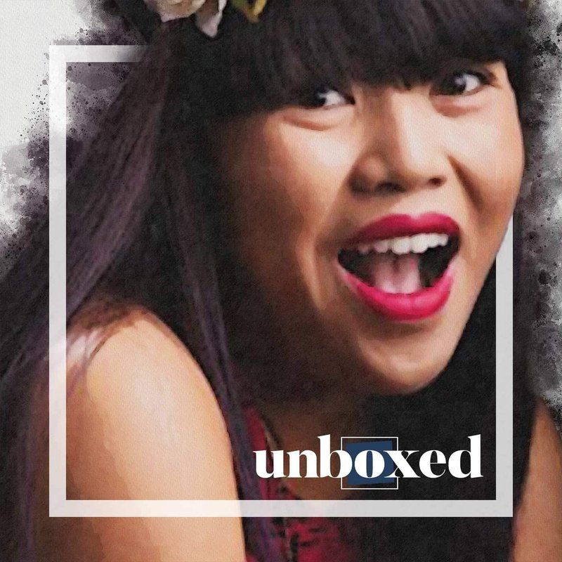 A person with long dark hair and vibrant makeup is smiling widely, reminiscent of a scene from Carol Cabreras student plays. The image has a stylized, painted effect, and the word unboxed is overlaid in the bottom right corner.