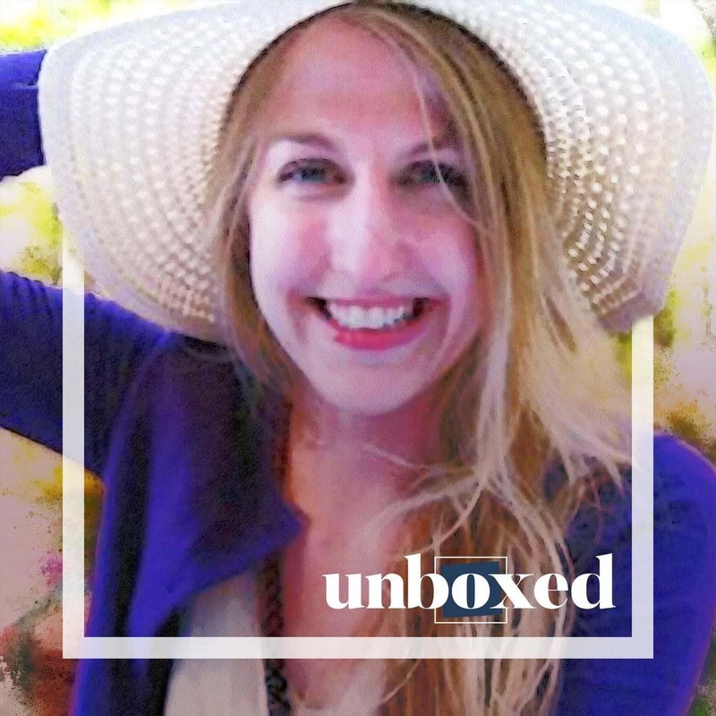 A person with long hair smiling, wearing a large, light-colored sun hat and a purple top. The background is blurred with warm colors, while unboxed graces the foreground. This vibrant image celebrates diversity and self-expression, reminiscent of the spirited essence of Julie Ruble.