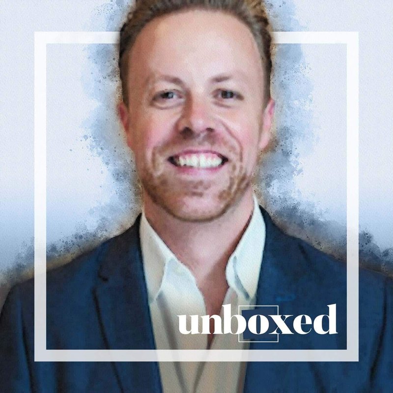 A person with short hair and a beard smiles at the camera, wearing a dark jacket over a light shirt that suggests professionalism. The image has a soft, artistic filter applied with the word unboxed in white text in the lower right corner, capturing the essence of creative work.