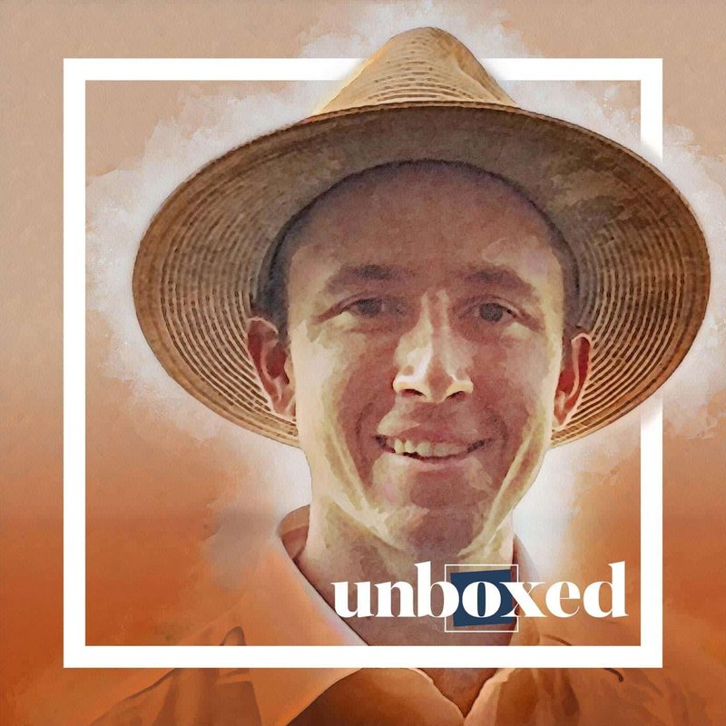 A man wearing a wide-brimmed straw hat smiles at the camera. The background is a soft gradient of orange hues, with unboxed overlaid at the bottom. A white square frame surrounds the image, capturing Shane Duenows approach to parallel prototyping in a visually captivating way.