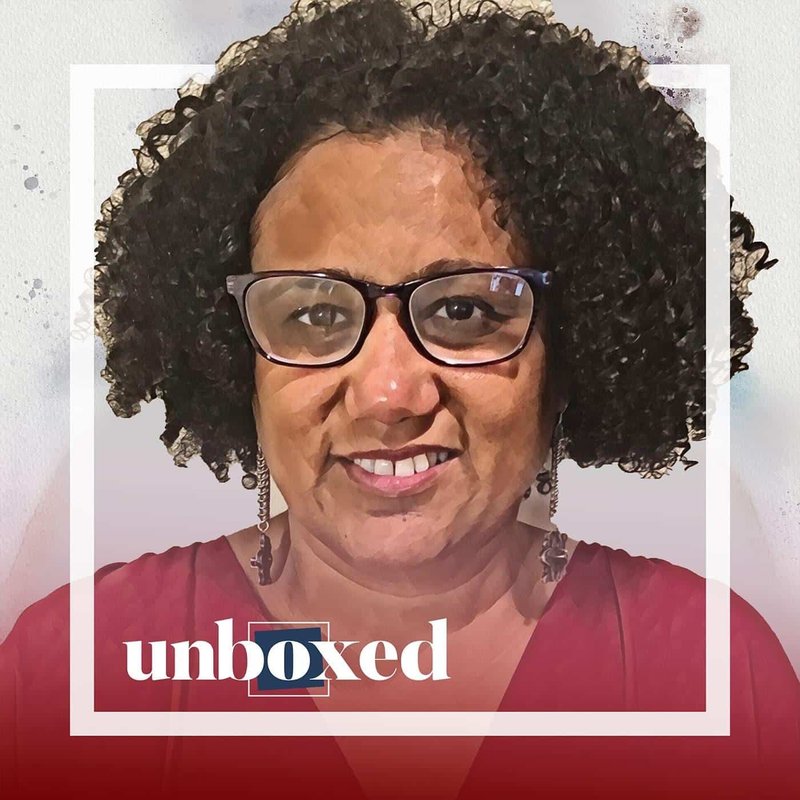 A woman with curly hair and glasses smiles warmly. She wears dangling earrings and a red top as if ready to dismantle barriers. The word unboxed appears at the bottom, echoing her spirit of breaking free from limitations.