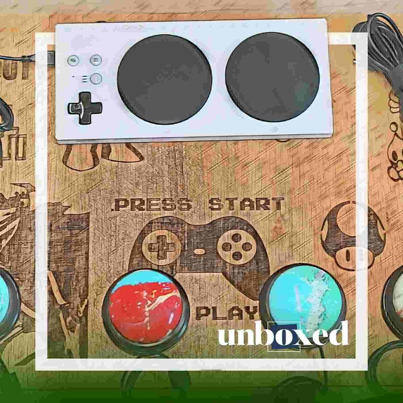 A gaming setup with a controller featuring a joystick and large round buttons on a wooden board, adorned with a gamepad graphic and the words PRESS START PLAY. This AbleGamerz project scene is framed with the word unboxed in the bottom corner.
