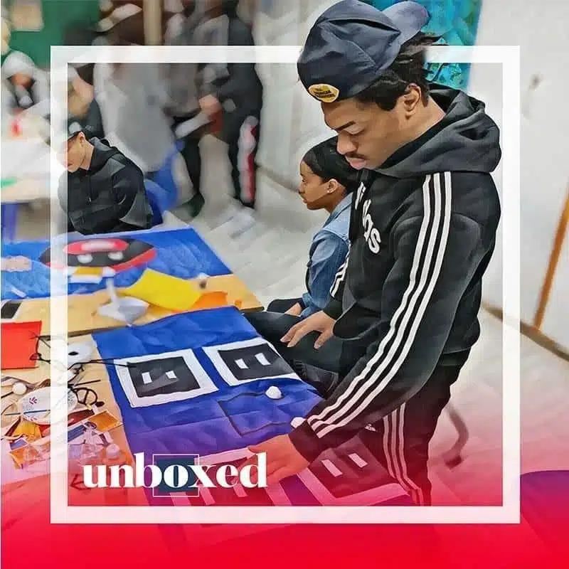A person in a black tracksuit and cap collaborates with others at a table, bringing vibrant geometric designs to life on the artwork. The word unboxed is displayed prominently in the foreground, as if teasing Episode 2 of their creative journey.