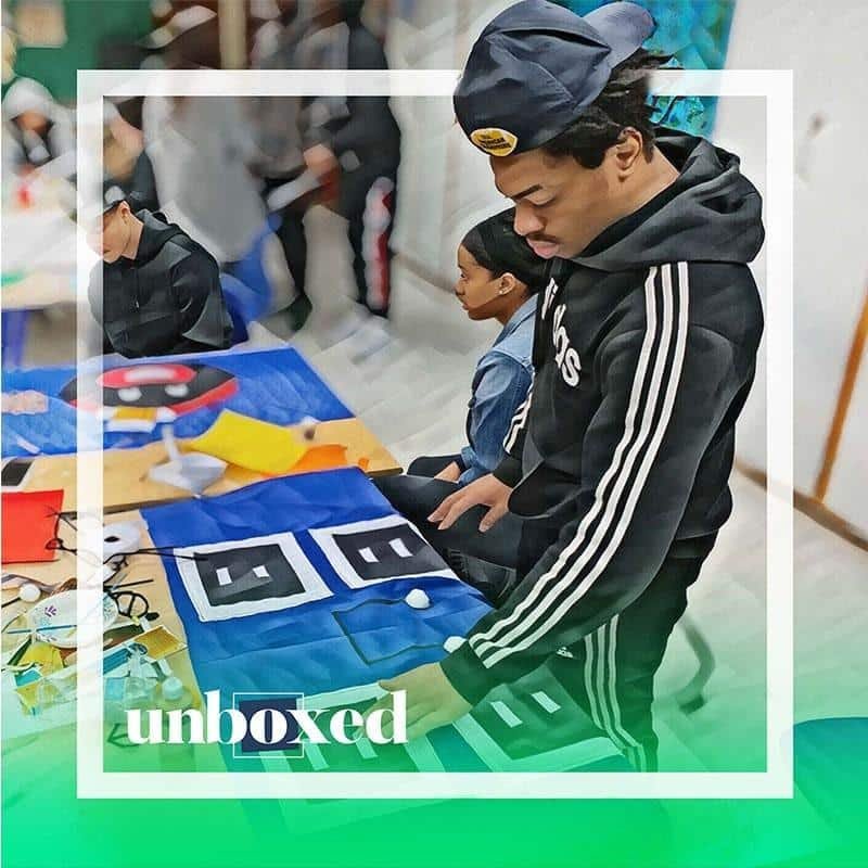 A person in a black tracksuit and cap is deeply focused on an art project, possibly quilting, on the table. Nearby, a young woman observes. The word unboxed hints at the creative spirit of Cheltenham PBL.