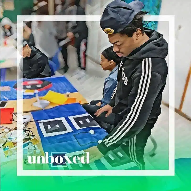 A person in a black tracksuit and cap is deeply focused on an art project, possibly quilting, on the table. Nearby, a young woman observes. The word unboxed hints at the creative spirit of Cheltenham PBL.