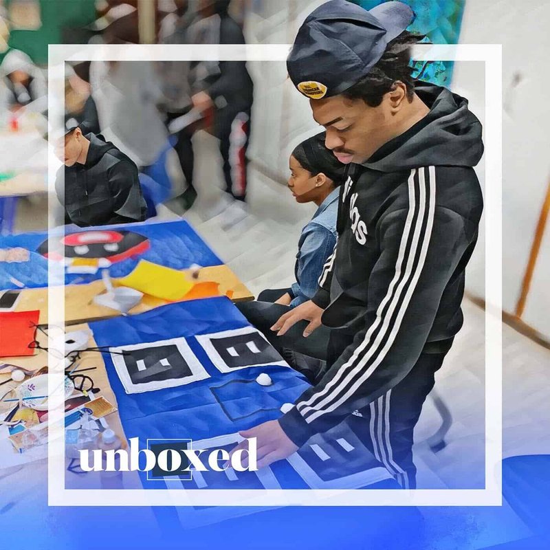 A person in a black hoodie and cap examines a patterned fabric piece on the table in Episode 4. Nearby, another sits surrounded by colorful materials, reminiscent of the creative chaos often seen in the Cheltenham PBL Experiment. The word unboxed is overlaid in the bottom left corner.