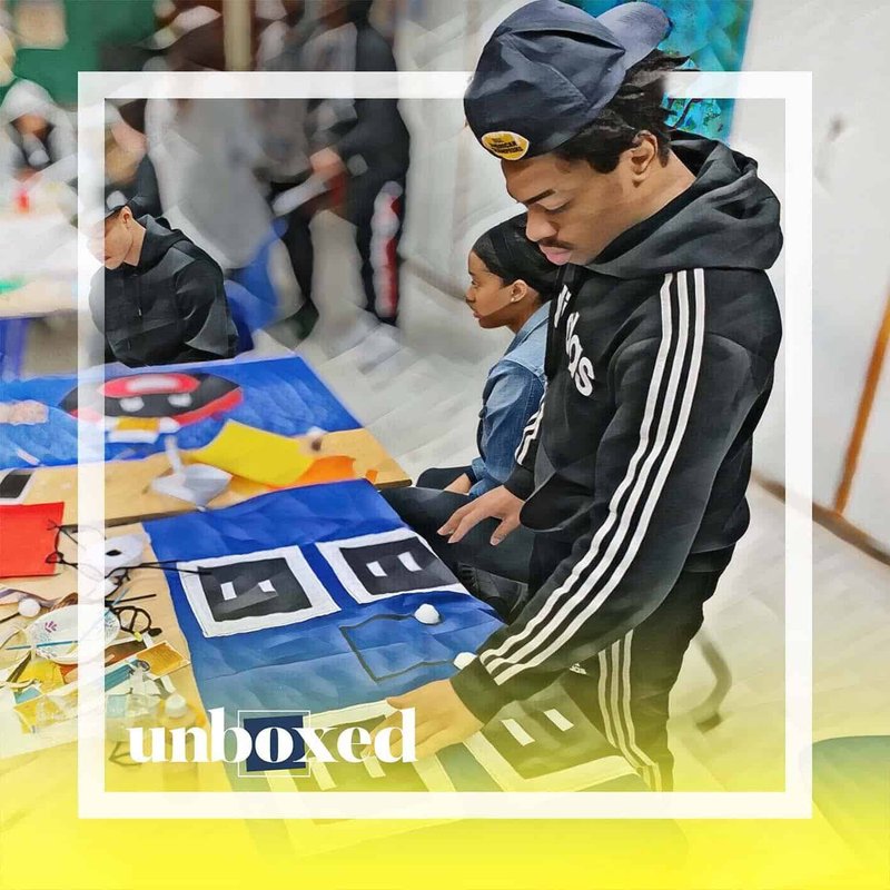 In March 2020, a person in a black hoodie and hat inspects a colorful quilt with abstract patterns. Nearby, another individual in a blue jacket sits observing. The scene, reminiscent of a Cheltenham art exhibit, is framed with the word unboxed at the bottom.
