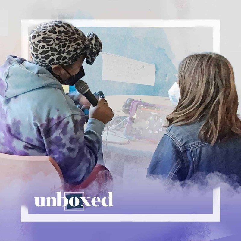 Two people sitting at a table, one holding a microphone, facing each other. The person on the left wears a leopard print headscarf and tie-dye hoodie, while the person on the right has long hair and a denim jacket. Text on the image reads unboxed, capturing the vibrant energy of Be Loud Studios Youth Radio Show in New Orleans.