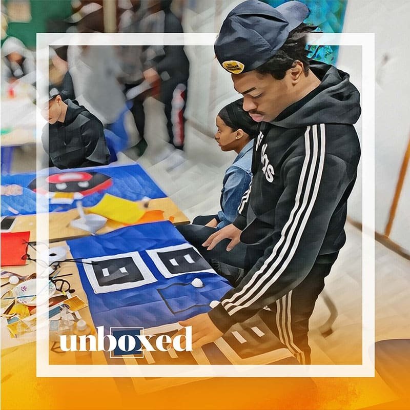 A young person in a black Adidas tracksuit and cap is focused on vibrant geometric artwork at a table. Nearby, another person sits engaged. The word unboxed appears with an orange gradient overlay, hinting at the creativity of the Cheltenham PBL Experiment.