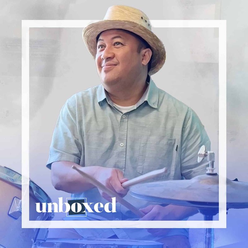 A person in a straw hat and blue shirt plays the drums, smiling as they look upwards, embodying the spirit of cultural celebration. The word unboxed is visible at the bottom left. The light background seamlessly blends with their attire.