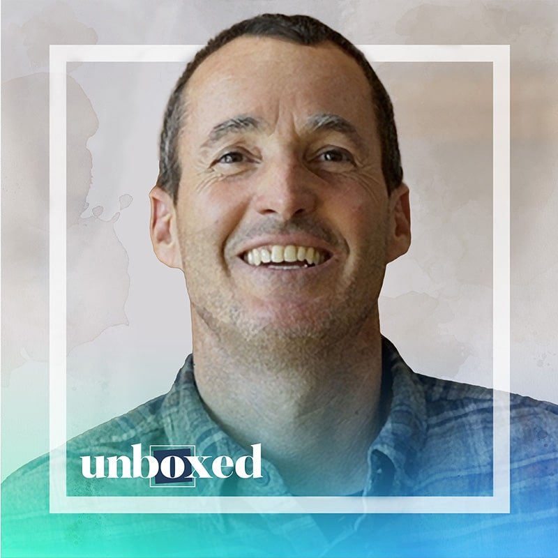 Smiling person wearing a plaid shirt within a square frame. The word unboxed is overlayed at the bottom in stylized text against a gradient background, celebrating John Santos latest design project launch.