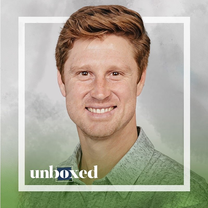A person with light brown hair smiling, wearing a gray button-up shirt. The image, reminiscent of an inspiring teachers PBL workshop, is framed with a white border and features the word “unboxed” in the bottom left corner. The background is a mix of gray and green tones.