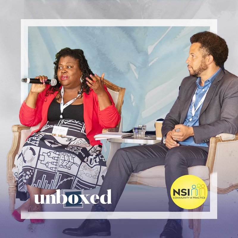 Dr. Brandi Hinnant-Crawford, in a red blazer, holds the microphone, gesturing passionately on stage during the Gates CoP NSI discussion. Dr. Luke Wood, clad in a blue shirt and blazer, listens intently beside her against a backdrop featuring the NSIM logo and unboxed text.