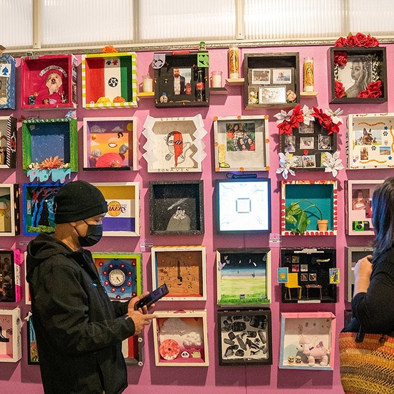 People stroll through a curated art exhibit featuring colorful shadow boxes on a pink wall. On the left, a person wearing a black mask holds up their phone, capturing the moment, while another gazes in admiration. The boxes celebrate diverse artworks and decorations, showcasing vibrant culture.
