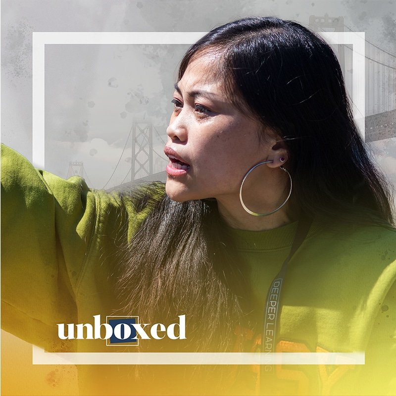 A person with long hair and hoop earrings gestures passionately like rapper Ruby Ibarra while wearing a green sweater. The image includes stylized elements, such as a faded bridge in the background, with the word unboxed overlaid at the bottom.