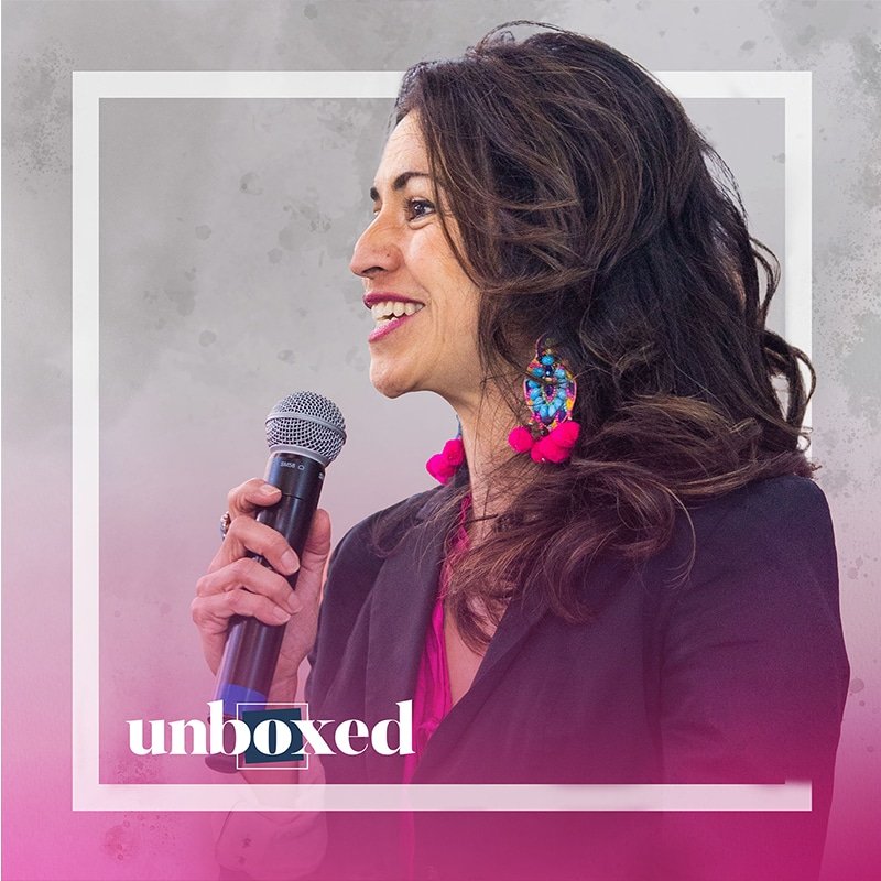 A woman with long hair, wearing colorful earrings and a pink outfit, is speaking into a microphone. She is framed by a white square with unboxed in white and pink at the bottom, echoing Rochelle Gutiérrezs vision of rehumanizing mathematics through inclusive dialogues.