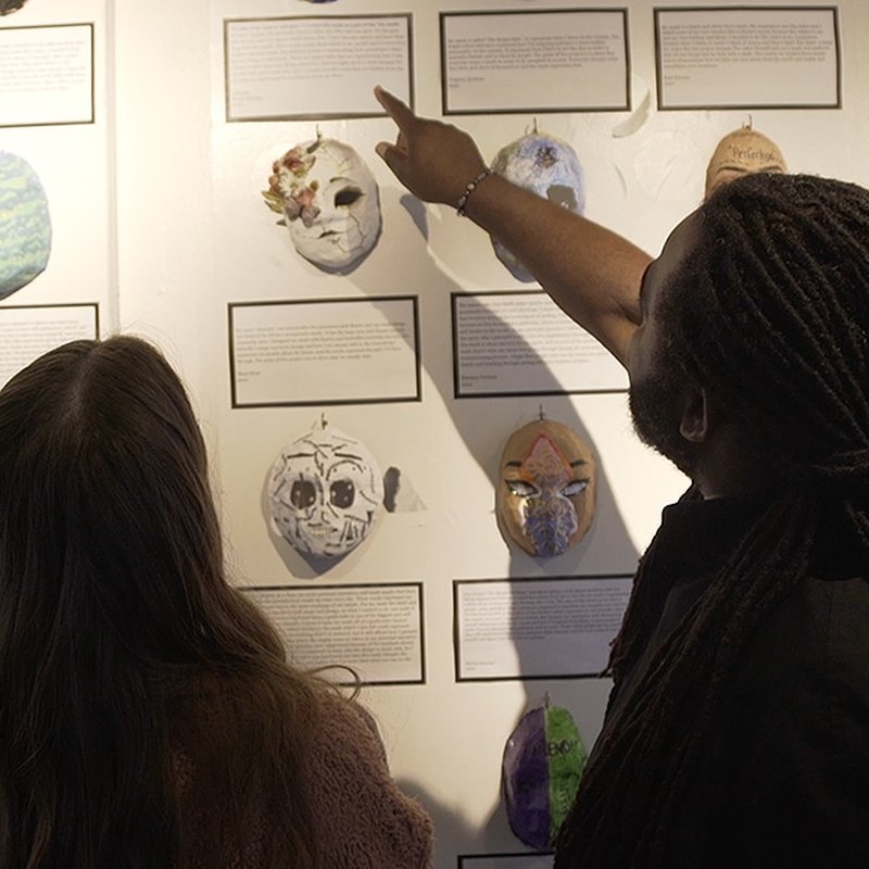 Two people examine a wall display of decorated masks and accompanying text, delving into a fascinating project. One person points towards a mask with their finger. The masks, showcasing skull and floral patterns, are framed by descriptive labels that enhance the social emotional learning experience.