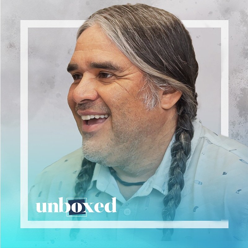A smiling man with long braided hair and a beard wears a light-colored shirt against a backdrop featuring the word unboxed. The blue gradient overlay perfectly captures the essence of an innovative project.