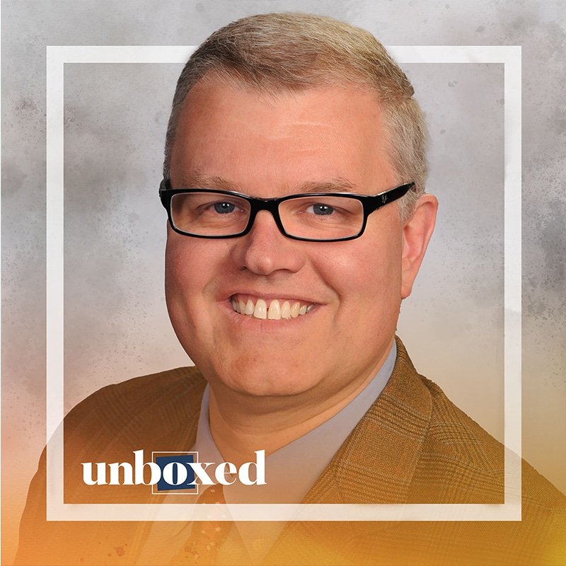 A person wearing glasses and a brown suit, embodying leadership, smiles at the camera in a photo with the word unboxed at the bottom. The image, evoking deeper learning, features a softly textured background.