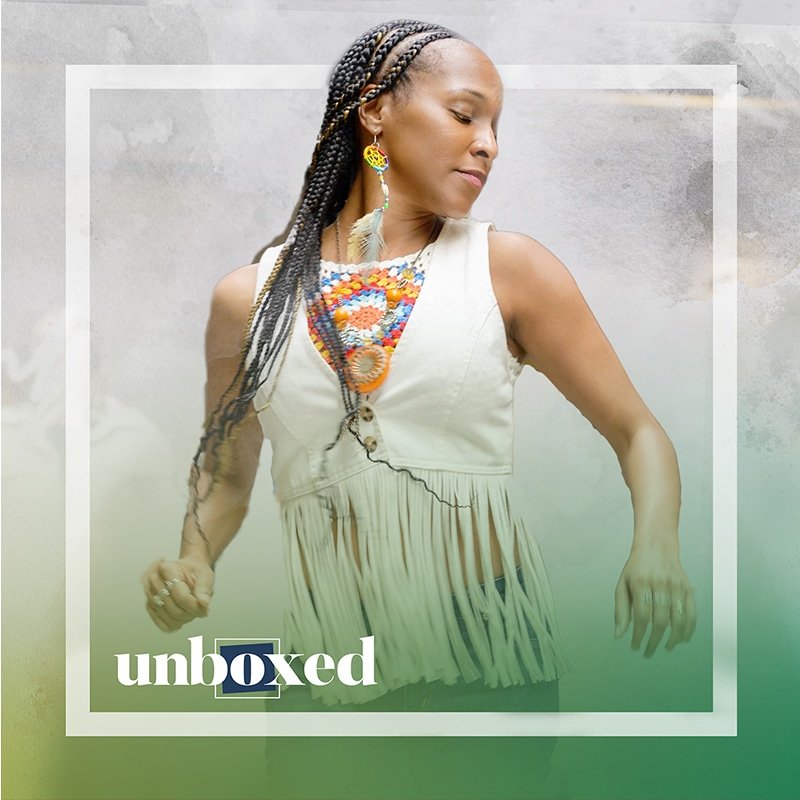 A woman with braided hair, reminiscent of Rodeo Days, wears a white fringed top and colorful beaded necklace. She stands with her eyes closed against a gray background, overlaid with the word unboxed at the bottom.