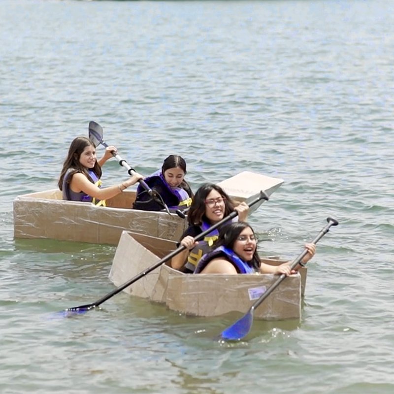 Four people in cardboard boats paddle across a serene lake, each wearing a life jacket and clearly enjoying the sunny day. This creative exhibition showcases simple materials, aligning with principles of project-based learning (PBL) and equity, as everyone participates equally in the fun.