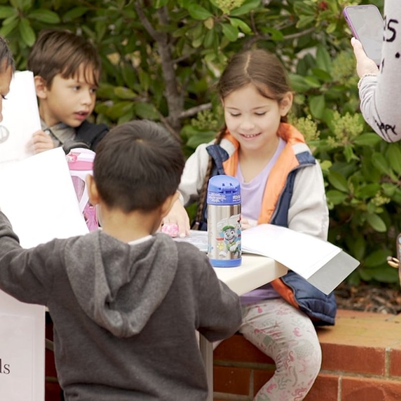 Children sit around a small outdoor table, engaged in collaborative design. One child holds a tablet while others interact enthusiastically. A water bottle sits on the table, surrounded by lively bushes in the background, capturing the essence of project-based learning in action.