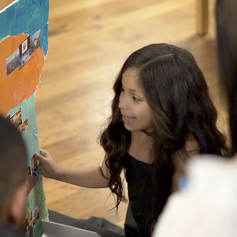 A young girl with long, wavy hair sits on the wooden floor, intently examining a colorful, personalized poster filled with photos. In the background, blurred figures suggest a lively exhibition space where each display captures her full attention.