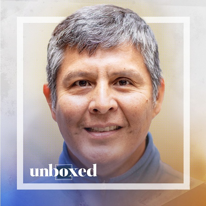 A smiling person with short gray hair, possibly Peter Deswood, is seen in a framed portrait. The image has a subtle blue and yellow gradient background with the word unboxed in the lower left corner, capturing a personal connection that resonates with Navajo students.