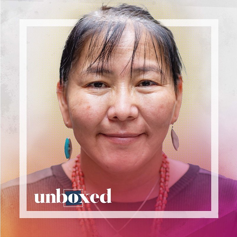 A person with short, dark hair and earrings is smiling at the camera, radiating a sense of identity. They are wearing a pink top and a beaded necklace. The image has a colorful border with unboxed in the bottom left corner, capturing Bernita Bedah’s vibrant spirit.