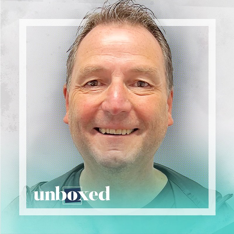 A smiling man in a black shirt and jacket is framed by a white square border with the text unboxed in the bottom left corner, subtly reminding us that work matters. The background features a soft gray and teal gradient.