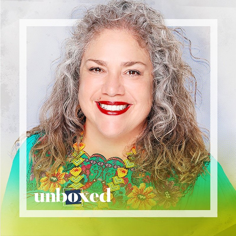 Smiling woman with long, curly gray hair, wearing a vibrant floral necklace and a teal blouse. She appears confident and joyful, embodying a heartset of positivity. The word unboxed is in the corner, partially obscured by a gradient.
